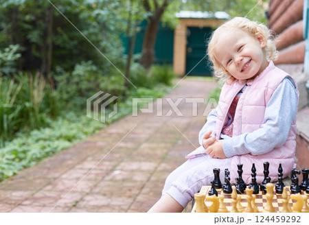 toddler girl smiles at the camera while sitting next to chess on the street in the garden. toddler girl smiles at the camera while sitting next to chess on the street in the garden. 124459292