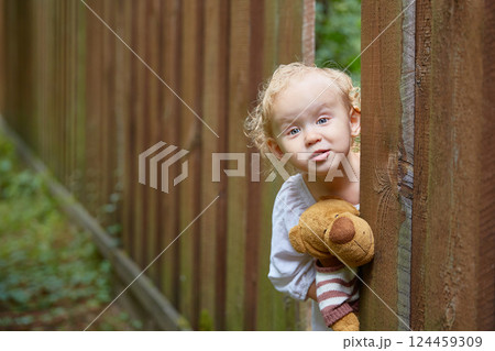 girl with a teddy bear in her hands looks out through a hole in a wooden fence. Copy space. 124459309