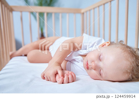 A cute baby in white clothes sleeps in a wooden crib in a bright, spacious room. 124459914