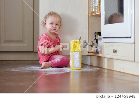 Playful laughing little baby girl smeared in flour sits on the kitchen floor. 124459949