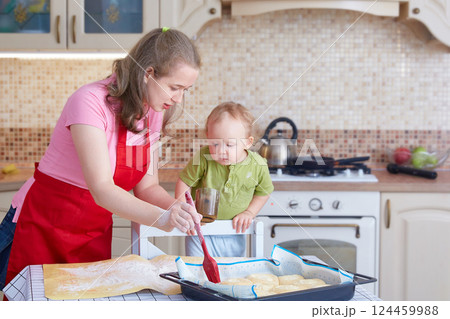 A young woman teaches a child to cook pastries. Greases the pies in the baking tray with a cooking brush. 124459988