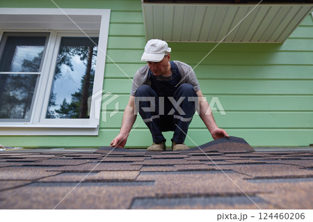 Male worker lays a sheet of soft flexible bitumen shingles on the roof of a country house. 124460206