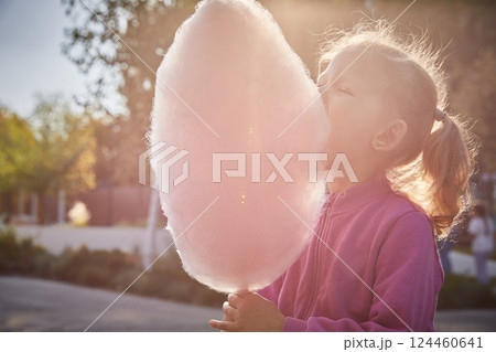 Cute curly-haired little girl eats cotton candy on a sunny summer evening in the park. Close-up. 124460641