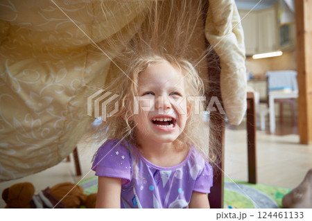 A preschooler girl laughs merrily sitting under a blanket in a shelter made at home. Electrified hair stands up. A preschooler girl laughs merrily sitting under a blanket in a shelter made at home. Electrified hair stands up. 124461133