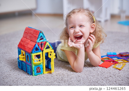 Portrait of a laughing girl on the carpet of the house next to a toy house made of magnets. Portrait of a laughing girl on the carpet of the house next to a toy house made of magnets. 124461265