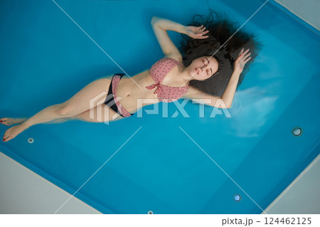 A young woman enjoys relaxing in a swimming pool or a bath with salt water. A brunette with loose hair is relaxing in a spa salon. Top view. Copy space. 124462125