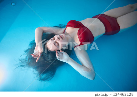 A young woman is swimming relaxed in a small pool, the blue background is visible through the clear water illuminated by light, dark hair, burgundy swimsuit, a day of relaxation at the spa. A young woman is swimming relaxed in a small pool, the blue background is visible through the clear water illuminated by light, dark hair, burgundy swimsuit, a day of relaxation at the spa. 124462129
