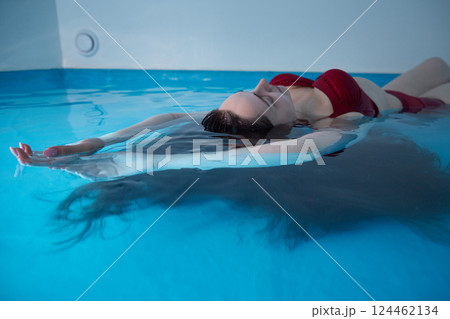 A young woman is swimming relaxed in a small pool, the blue background is visible through the clear water illuminated by light, dark hair, burgundy swimsuit, a day of relaxation at the spa. 124462134