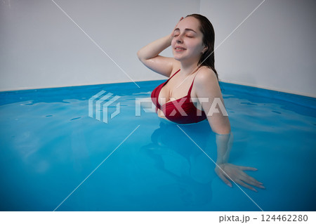 A young woman is swimming relaxed in a small pool, the blue background is visible through the clear water illuminated by light, dark hair, burgundy swimsuit, a day of relaxation at the spa. 124462280