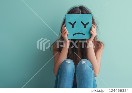 A young woman sitting against a teal wall holds a sad blue face card in front of her face, hiding her emotions 124466324