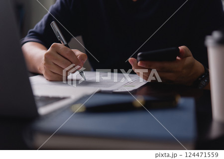 man writing content or a script on a notepad with a pen while holding a phone, highlighting multitasking in work, journalism, or education using a notebook and laptop. man writing content or a script on a notepad with a pen while holding a phone, highlighting multitasking in work, journalism, or education using a notebook and laptop. 124471559