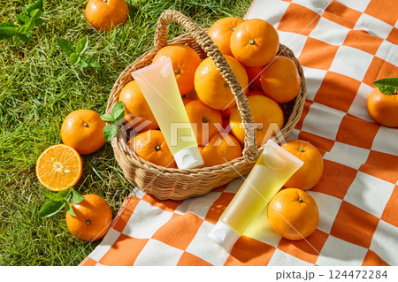 Top view of tubes of cream placed among fresh oranges in a handwoven bamboo basket. Oranges are scattered around on the grass and checkered picnic mat. Mockup plastic tube for design. 124472284