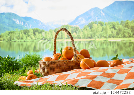 Lawn with green bushes next to the river with a clear lake reflecting the mountain view. Wicker basket containing Citrus Burst Oranges. Ripe oranges with rough peel placed on checkered carpet 124472288