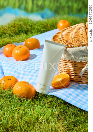 Oranges placed on checkered carpet and on lawn with upright grass blades. White cream tube placed between oranges and handwoven wicker basket. vertical shot for advertising, outdoor picnic concept 124472299