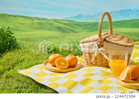 Fresh oranges cut in half and into wedges placed on round wooden plates, presented on white and yellow checkered squares. Wicker baskets with handles and lace trim set against a backdrop of lawns 124472304