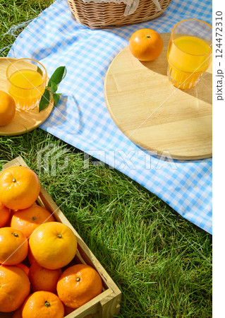 A glass of orange juice sits on a smooth, round wooden cutting board. A small blue and white checkered cloth is spread over a soft lawn. A light brown wooden crate is filled with ripe oranges. 124472310