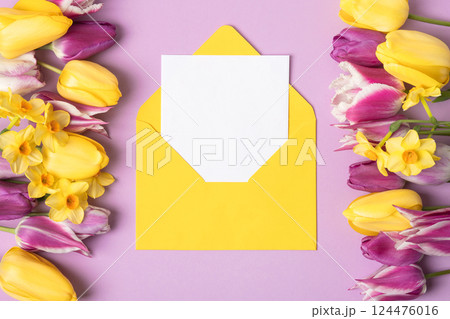 Yellow envelope with clean white sheet of paper for your text and spring flowers purple yellow tulips and yellow daffodils on lilac background. Invitation to the holidays. Flat lay. 124476016