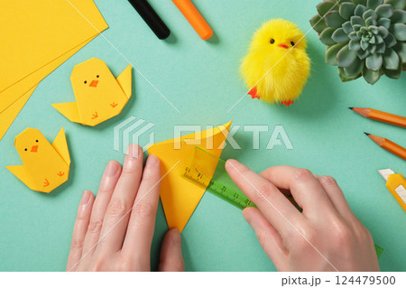 Making a simple Easter origami handicraft. Hands fold a square yellow sheet in half with a ruler. Around ready-made crafts chickens and tools for crafts 124479500