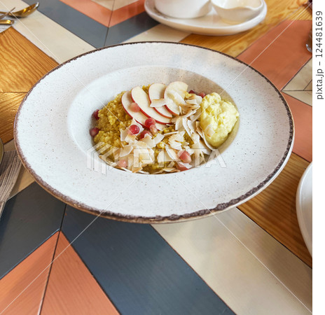 Nutritious breakfast bowl featuring millet topped with fresh apple slices, pomegranate seeds, and coconut flakes, gluten free Presented on a colorful wooden table setting in a cafe Nutritious breakfast bowl featuring millet topped with fresh apple slices, pomegranate seeds, and coconut flakes, gluten free Presented on a colorful wooden table setting in a cafe 124481639