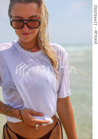 A woman in a wet white shirt and sunglasses stands on the beach 124482082