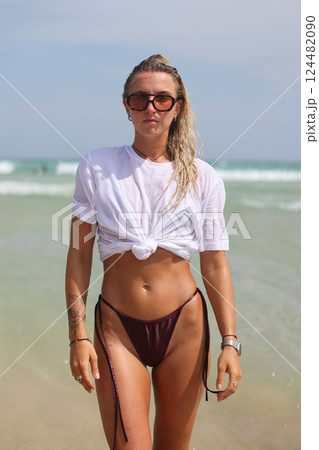 A woman dressed in a bikini and wearing sunglasses is walking along the beach A woman dressed in a bikini and wearing sunglasses is walking along the beach 124482090