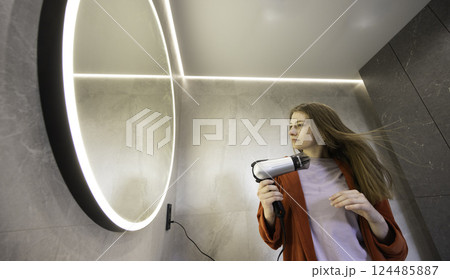 Young woman in a red robe drying her long hair with a hairdryer in a modern bathroom with a glowing round mirror. Young woman in a red robe drying her long hair with a hairdryer in a modern bathroom with a glowing round mirror. 124485887