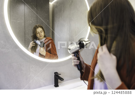 Young woman in a red robe drying her hair with a hairdryer while looking at herself in the round illuminated bathroom mirror. Young woman in a red robe drying her hair with a hairdryer while looking at herself in the round illuminated bathroom mirror. 124485888