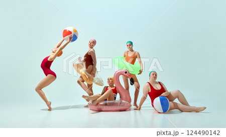 Five young women in multicolored swimsuit and caps posing with beach essentials, balls and inflatable toys against light blue background 124490142