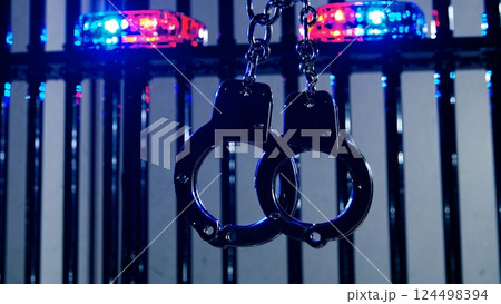 Metal handcuffs dangling from prison cell bars, backlit by pulsing emergency lights, signaling law enforcement detention and criminal restraint 124498394