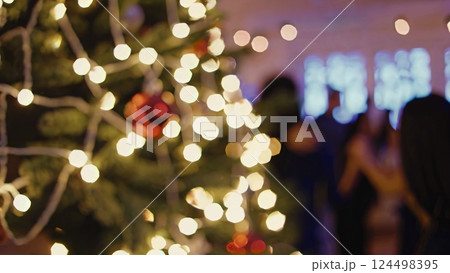 Glowing holiday lights highlighting decorated evergreen tree, festive blurred guests celebrating winter season with soft bokeh background. Warm christmas lights illuminate a decorated tree 124498395