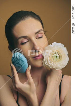 Woman using skincare device while holding a flower with closed eyes 124499055