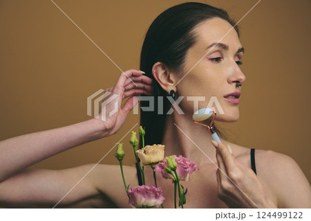 Woman using jade roller on her face while holding flowers in a serene setting Woman using jade roller on her face while holding flowers in a serene setting 124499122