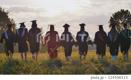 Group of graduates walking away in field at sunset holding hands, celebrating their academic achievement and friendship Group of graduates walking away in field at sunset holding hands, celebrating their academic achievement and friendship 124506513