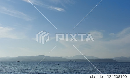 This photo captures a serene coastal scene. The image shows a vast, calm ocean extending into the horizon under a bright blue sky filled with scattered, fluffy white clouds. On the left side of the ph This photo captures a serene coastal scene. The image shows a vast, calm ocean extending into the horizon under a bright blue sky filled with scattered, fluffy white clouds. On the left side of the ph 124508190