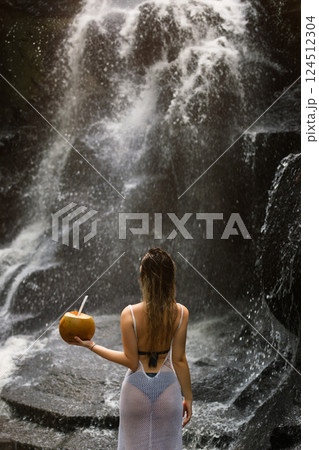 Beautiful sexy woman stands facing a waterfall in Bali and holds a coconut in her hand 124512304