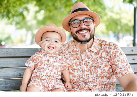 Stylish father and baby wearing matching outfits, sitting on a park bench smiling 124512978