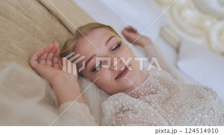 Elegant bride relaxing on bed, wearing white wedding gown with pearl accents, soft veil draping, capturing serene wedding morning moment 124514910