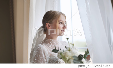 White clad bride holding wedding bouquet, gazing through window with soft, romantic expression 124514967