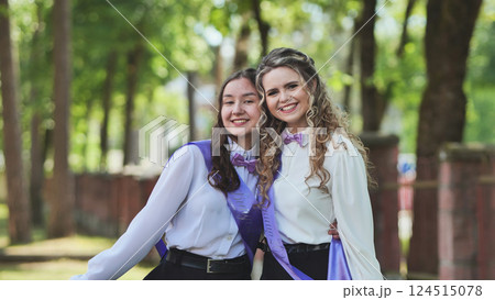 Two young women, graduates, hugging on final school day, radiating happiness in lush green park. Celebrating achievement, looking forward to bright future 124515078