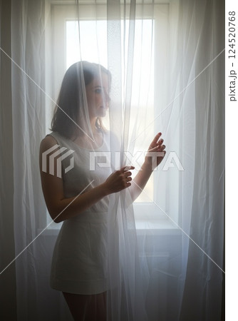 European brunette woman holding sheer white curtain with soft light in linen interior 124520768