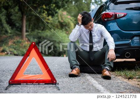 In the face of car trouble, a young businessman calmly changes the wheel of a broken car, while a worried mature man sits beside him. A red triangle sign warns drivers to proceed with caution. 124521778