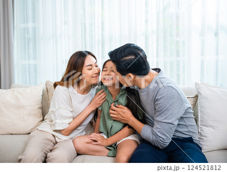 Family Day. Happy Asian father and mother kissing daughter in a living room at home, family together enjoy with love, Parents kiss cheek child girl while sitting on couch or sofa together 124521812