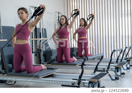 Three women in pink sportswear doing pilates exercises with a reformer bed.  124522690