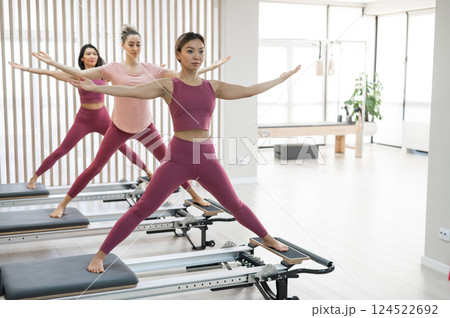 Three Asian women in pink sportswear doing pilates exercises with a reformer bed.  124522692