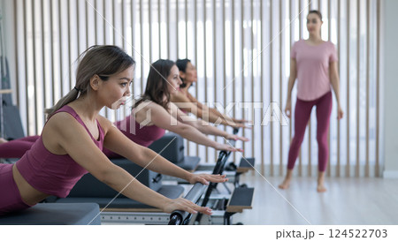 Three women in pink sportswear doing pilates exercises with a reformer bed.  124522703