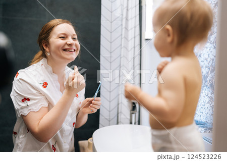 Joyful mother and toddler daughter having fun in bathroom while brushing teeth, toddler girl touching mother nose with finger. Concept of scene curiosity and innocence in bathroom setting. Joyful mother and toddler daughter having fun in bathroom while brushing teeth, toddler girl touching mother nose with finger. Concept of scene curiosity and innocence in bathroom setting. 124523226