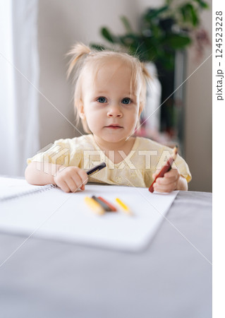 Vertical portrait of beautiful child girl drawing with colored pencils on paper pad sitting at table in living room with bright interior. Concept of childhood development, early learning, education. 124523228