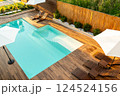 Modern turquoise swimming pool with wooden deck, lounge chairs, and umbrellas in a stylish backyard 124524156