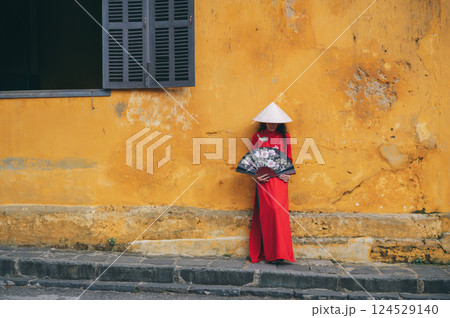 Asian woman tourist in traditional vietnamese non la hat and red dress with a fan on an old town street in Hoi An in Vietnam in Asia 124529140