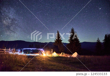 Blurred silhouettes of hikers resting around campfire, surrounded by illuminated tents under stunning starry sky. Milky Way stretches above, creating magical atmosphere in mountain wilderness. Blurred silhouettes of hikers resting around campfire, surrounded by illuminated tents under stunning starry sky. Milky Way stretches above, creating magical atmosphere in mountain wilderness. 124533133